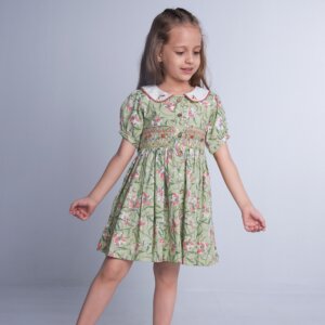 In a green floral smock dress, a child stands with hands straight, the collar adorned with delicate hand-embroidered details