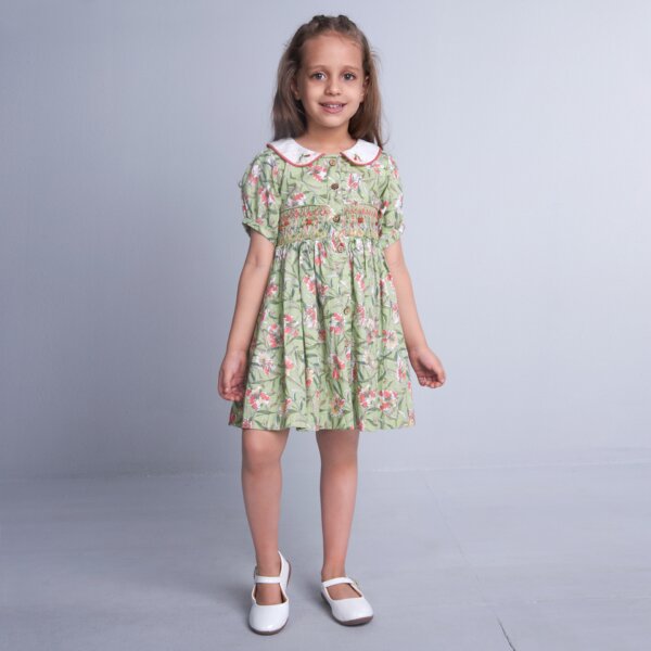 A child stands in a green floral smock dress, adorned with delicate hand-embroidered details on the collar