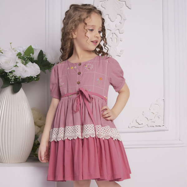 A girl in pink embroidered ombre dress with lace detail, tie up sash, and wood button fastening
