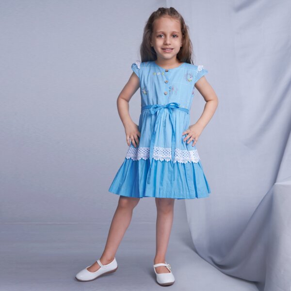 With hands on her hips, a young girl showcases a blue ombre dress adorned with delicate embroidery and lace details, featuring a tie-up sash