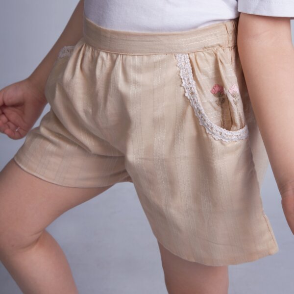 Posing from the side, a girl wears a tan hand embroidered shorts with dual side pockets and lace trims