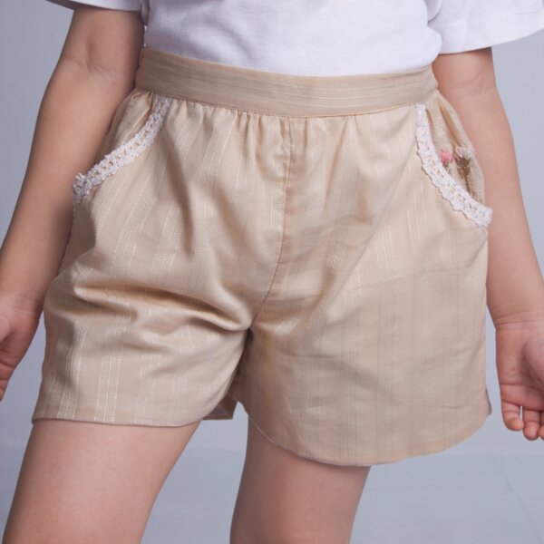 A little girl in cotton tan shorts, designed with a delicate touch of lace trims and hand embroidery on the pocket