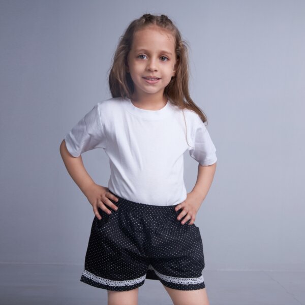With a hand on her hips, a girl poses in black dot printed shorts adorned with white lace trims and dual pockets