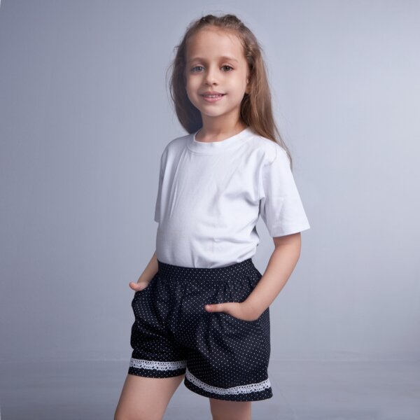Hand inserted in the pocket, a little girl stands in black dot printed shorts with white lace trims
