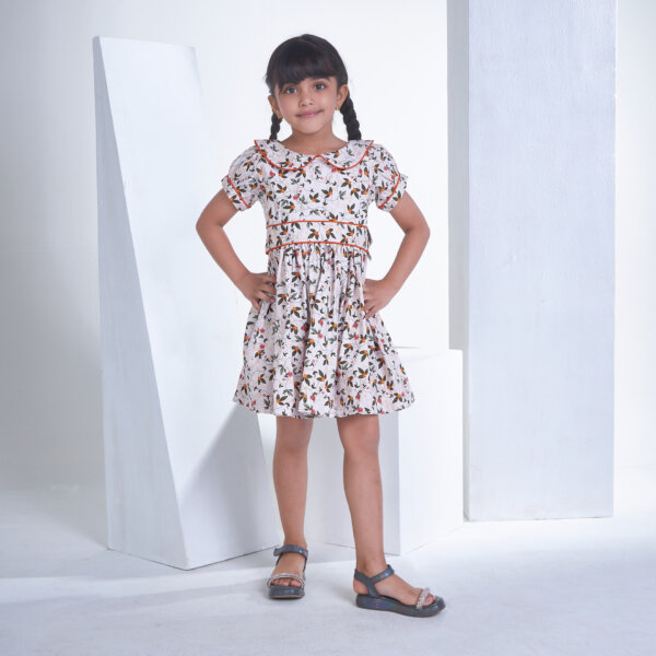 With hands on hips, a little girl stands in an ivory floral printed collared dress with an adjustable belt at the waist