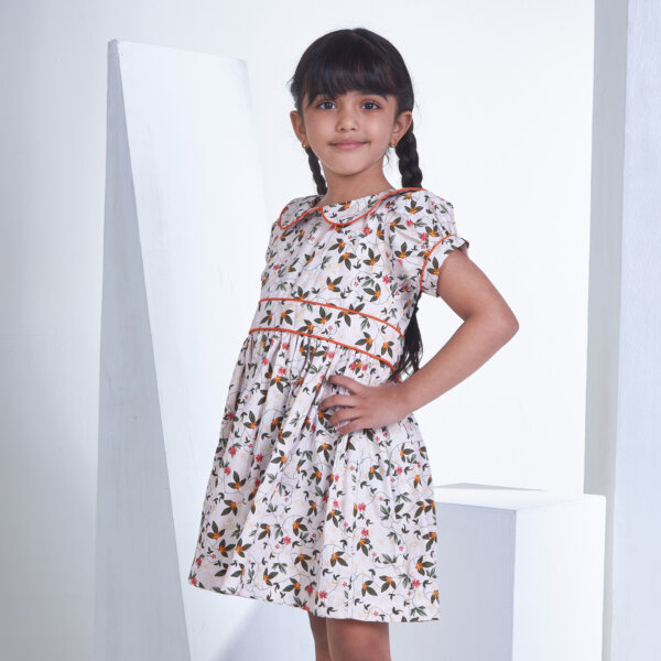 From the side, a little girl in an ivory floral printed collared dress with an adjustable belt