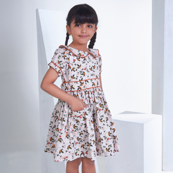 Hand inserted in the pocket, a little girl stands in an ivory floral printed peter pan collared dress with an adjustable belt