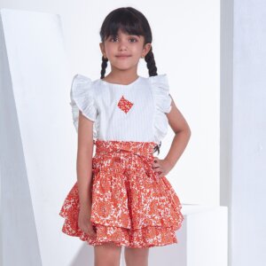 A little girl in an ivory embroidered ruffled sleeveless blouse and orange floral printed high waist skirt