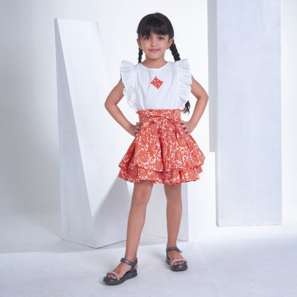 With hands on hips, a little girl stands in an ivory embroidered ruffled sleeveless blouse and orange floral printed high waist skirt
