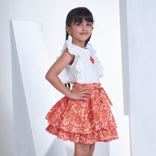In side view, a girl wears an ivory embroidered ruffled sleeveless blouse and orange floral printed high waist skirt