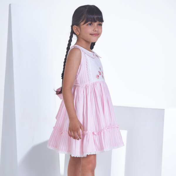 Posing from the side, a little one wears a pink striped embroidered collared dress adorned with ruffled hem