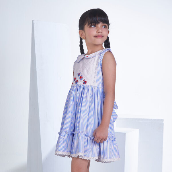 Posing from the side, a little one wears a blue striped embroidered collared dress adorned with ruffled hem